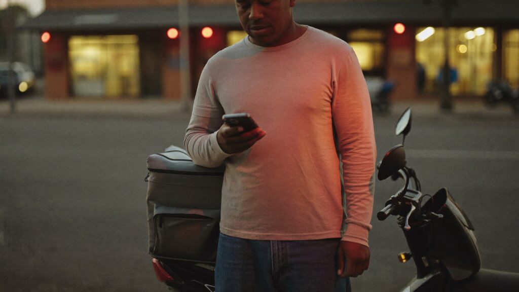 Entregador de aplicativo confere o celular ao lado da moto, em uma rua comercial no fim da tarde