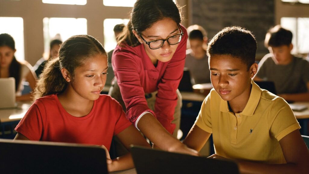 Professora ajuda dois estudantes com um notebook em uma sala de aula pública